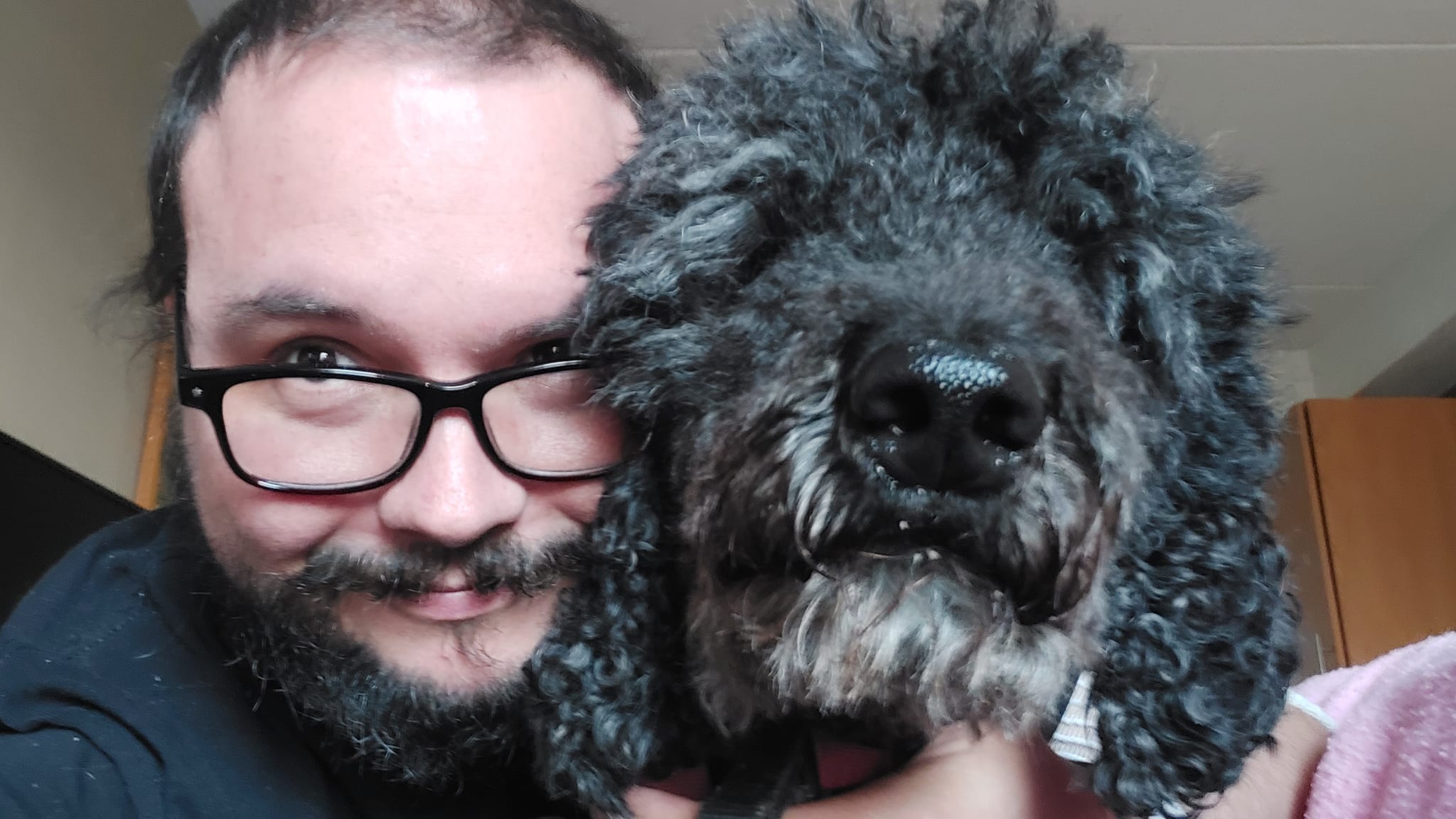 A man short hair and bushy facial hair hugs a shaggy black standard poodle, you can tell where her eyes are, but cannot see them under the shag